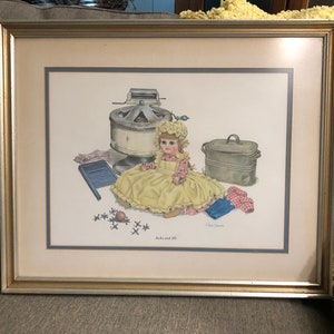 May include: A framed print of a vintage illustration of a doll dressed in a yellow dress and a pink and white bonnet. The doll is sitting on a floor with a variety of vintage household items, including a wringer washer, a washboard, and a metal container. The print is titled "Jack and Jill" and signed "Glen Easton".