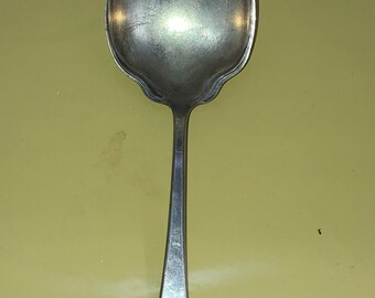 May include: A silver spoon with an ornate design on the handle. The spoon is on a yellow surface.