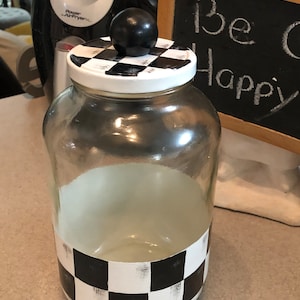 May include: A clear glass jar with a white lid featuring a black and white checkerboard pattern. The jar has a black and white checkerboard design around the base and a black sphere on top. A small chalkboard with the words "Be Happy" is in the background.
