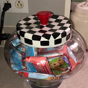 May include: A clear glass jar with a black and white checkered lid and a red knob. The jar is filled with individually wrapped granola bars.