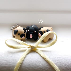 May include: Three small pug figurines in tan and black with pink tongues and black eyes. One has a silver loop for a keychain. A yellow ribbon is tied in a bow in the foreground. The pugs are on a white surface.
