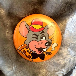 Puede incluir: Botón redondo naranja con una ilustración de un ratón de dibujos animados. El ratón es gris con detalles rosas, lleva un sombrero rojo y una pajarita negra, guiñando un ojo y sosteniendo pizza. El botón tiene un acabado brillante.