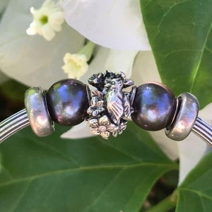 May include: A silver charm bracelet featuring a bird and flower charm, two dark grey beads, and two silver beads.