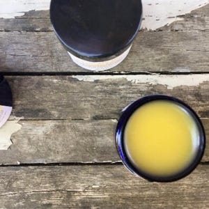 May include: Overhead shot of a wooden surface with three containers. Two are black with lids, and one is a small, round, blue-rimmed bowl filled with a yellow liquid. The wood has a weathered, distressed appearance.