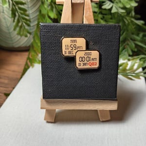 May include: A small wooden easel displays a black textured square with two rectangular pins. The pins are gold-colored with black text indicating dates and times: "1999 11:59pm 31 DEC" and "2000 00:01am 01 JAN QE2". Green foliage is visible in the background.