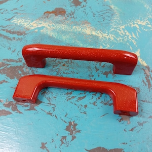 May include: Two red plastic cabinet door handles with a textured finish. The handles are different sizes.