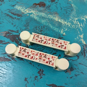May include: Two white ceramic drawer pulls with a floral design. The pulls have a rounded end and a flat rectangular center with a red and white floral pattern.