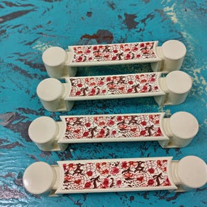 May include: Four white plastic drawer pulls with a floral design in shades of pink and brown. The pulls have a rounded knob on each end.