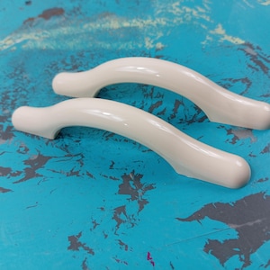 以下が含まれることがあります： オフホワイトのプラスチック製のキャビネットドアハンドル2個。ハンドルは湾曲しており、滑らかな仕上げになっています。