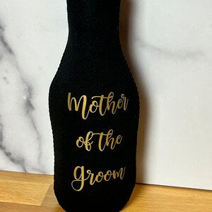 May include: Black bottle cooler with a gold zipper and the text "Mother of the Groom" in gold lettering.