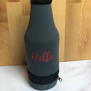 May include: Gray neoprene bottle cooler with a zipper and the name "Nellie" printed in red.