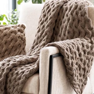 Puede incluir: Un sillón con asiento tapizado de color crema y estructura de metal negro. Una manta de punto marrón con un patrón de trenzas texturizado está colocada sobre el sillón, y un cojín cuadrado de punto marrón a juego descansa sobre el asiento.