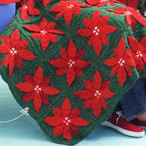 Pode incluir: Uma manta de crochê verde e vermelha com um padrão de poinsettia. A manta está drapeada sobre um banco de madeira vermelho e uma pessoa está sentada no banco.