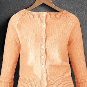 May include: A peach-coloured, button-down cardigan sweater hangs on a wooden hanger against a dark grey background. The sweater has long sleeves, ribbed cuffs, and a ribbed hem. The buttons are white.