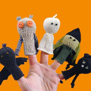 May include: A set of five knitted finger puppets featuring a black bat with wings, a beige owl with orange button eyes, a white ghost, a green-haired figure in a black hat, and a black cat with white button eyes, all displayed on fingers.
