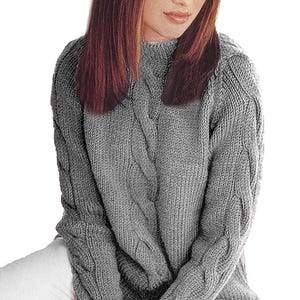 以下が含まれることがあります： ハイネックのグレーのケーブルニットセーター。袖と前面にテクスチャードケーブルデザインが施されています。モデルは白いパンツを着用しています。セーターは柔らかく暖かい素材で作られています。