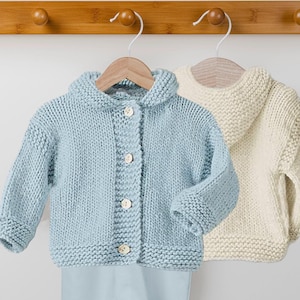 Peut inclure: Deux pulls en tricot pour bébé, un bleu clair et un crème, accrochés à des crochets en bois. Les deux pulls ont une capuche et une fermeture à boutons.