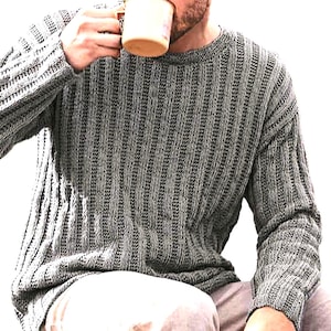 Puede incluir: Un suéter de punto gris con un diseño acanalado. El suéter tiene mangas largas y cuello redondo. La persona sostiene una taza de color marrón claro.