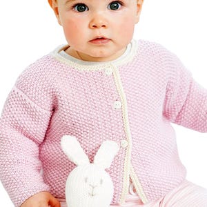Può includere: Un bambino indossa un cardigan rosa chiaro lavorato a maglia con finiture e bottoni color crema, abbinato a pantaloni a righe rosa. Il bambino tiene in mano un giocattolo a forma di coniglio bianco lavorato a maglia. Lo sfondo è bianco.