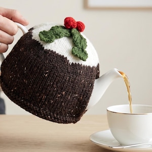 Peut inclure: Une théière en céramique blanche recouverte d'une housse tricotée marron foncé et blanche, ornée d'une feuille verte et de baies rouges sur le dessus, versant du thé dans une tasse et une soucoupe assorties avec un bord argenté et une cuillère.