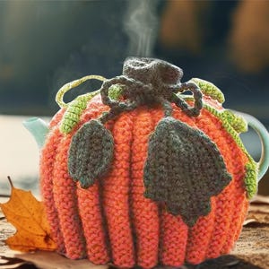 Op de afbeelding: Een turquoise theepot met een gebreide oranje pompoenhoes, groene bladeren en een donkergrijze stengel. Links staan een kopje thee en een lepel op een schoteltje.