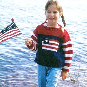 May include: A young girl wearing a blue, red, and white striped sweater with a red, white, and blue American flag design on the front. She is holding a small American flag in her right hand and is standing in shallow water with her feet bare.