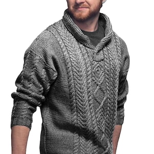May include: A grey cable-knit jumper with a shawl collar. The jumper features a textured pattern with diamond and chevron designs. The long-sleeved garment is made of a thick, warm material, suitable for cooler weather. The jumper has a button closure at the neckline.