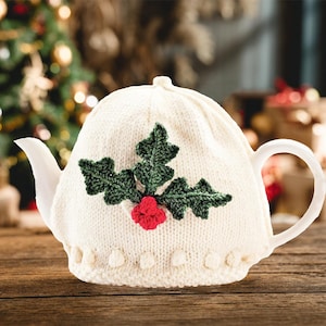 May include: A white ceramic teapot with a knitted cozy featuring a green holly leaf design and red berries. The cozy has a decorative border of small white bobbles. The teapot is on a wooden surface.
