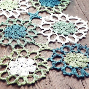 May include: A crocheted table runner made of white, green and blue yarn. The runner is made up of several individual crocheted circles that are joined together.