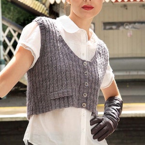 May include: A woman wearing a white blouse with a gray knitted vest and black leather gloves. The vest has a scoop neck and a button closure.