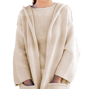 May include: A beige knit hooded cardigan featuring long sleeves, an open front design, and two large patch pockets at the bottom. The garment is crafted from a solid-coloured yarn with a consistent stockinette stitch pattern throughout the piece.