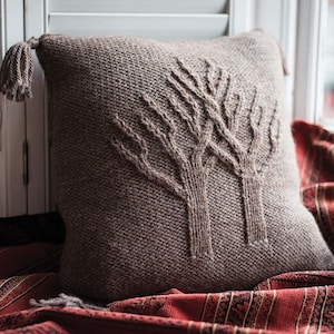 Peut inclure: Un coussin en tricot marron avec un motif tissé de deux arbres. Le coussin a un pompon sur un coin et est posé sur une couverture rayée rouge et noire.