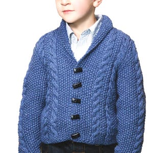 May include: A blue cable-knit cardigan featuring a shawl collar and four black toggle buttons down the front. The sweater is worn over a light blue and white pinstriped collared shirt, paired with dark blue denim jeans.