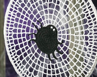 Può includere: Un ornamento circolare a forma di ragnatela lavorato all'uncinetto in bianco, con un ragno nero lavorato all'uncinetto con due piccoli occhi bianchi al centro. La ragnatela è realizzata con un filo bianco sottile e appesa a un filo bianco.