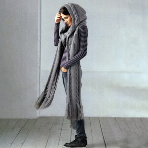 May include: A grey hooded scarf with a cable knit pattern and fringe. The scarf is worn by a person with a striped shirt and jeans.