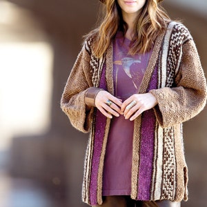 Può includere: Un cardigan marrone con strisce viola e bianche. Il cardigan ha un'apertura anteriore lunga ed è realizzato in un tessuto morbido e testurizzato.