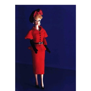 Op de afbeelding: Een rode jurk met een zwarte riem en zwarte handschoenen. De jurk heeft een strakke rok en een jasje met een brede kraag. De pop draagt een zwarte hoed met een rode strik.