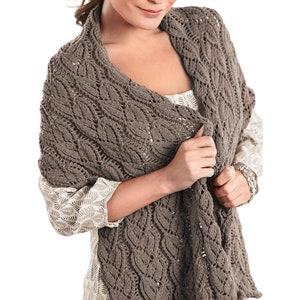 May include: A brown knitted shawl with a delicate cable pattern. The shawl is draped over a person's shoulders and is being held in place by one hand.
