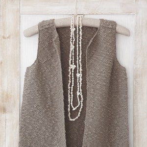 Könnte beinhalten: Eine braune, gestrickte, ärmellose Weste, die an einem Holzbügel hängt, mit einer weißen Muschelkette darüber.