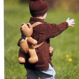 May include: A child's knitted brown sweater and hat set. The sweater features a kangaroo pocket with a teddy bear and a backpack with a teddy bear. The child is wearing blue jeans and brown boots.
