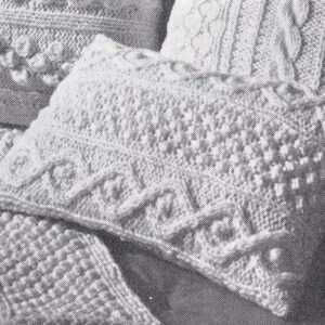May include: A close-up of a white knitted pillow with a textured pattern. The pillow is made with a cable stitch and has a raised design.