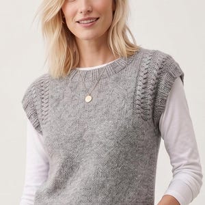 May include: A woman wearing a gray knitted vest over a white long-sleeved shirt. The vest has a round neckline and cable knit detailing on the shoulders. A gold pendant necklace is visible.