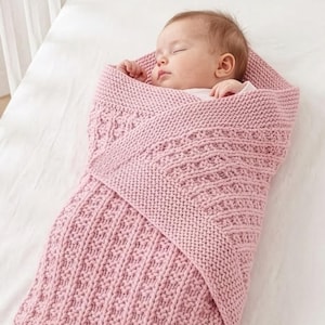 May include: A sleeping baby wrapped in a soft, pink knitted blanket inside a white crib. The blanket has a textured pattern. A mobile with star-shaped ornaments hangs above the crib.