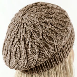 May include: A light brown knitted beret featuring a textured cable knit pattern that radiates from the center crown down to a ribbed brim. The hat is constructed with a soft yarn material and displays a consistent, intricate stitch design throughout.