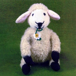 May include: A white knitted plush toy sheep with black hooves and a green stem with a white flower in its mouth. The sheep is sitting on a green surface.