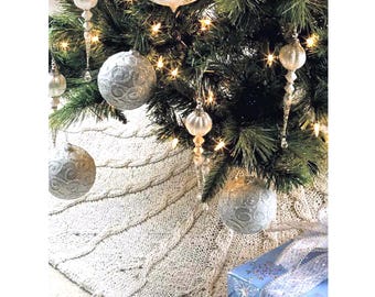 Könnte beinhalten: Ein weißer gestrickter Christbaumrock mit einem Zopfmuster. Der Rock ist mit silbernen Kugeln und einer blauen Geschenkbox mit silbernem Band verziert.