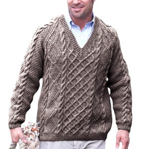 Könnte beinhalten: Ein brauner Zopfstrickpullover mit V-Ausschnitt. Der Pullover hat komplizierte Zopfmuster auf Brust und Ärmeln. Die Ärmel haben ein dickes Zopfmuster. Darunter wird ein hellblaues Hemd getragen.