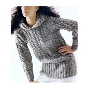Peut inclure: Un pull à capuche gris avec un motif en tricot torsadé. Le pull a des manches longues et un ourlet côtelé. Le modèle porte un pantalon blanc. Le pull est fait d'une matière douce et chaude.