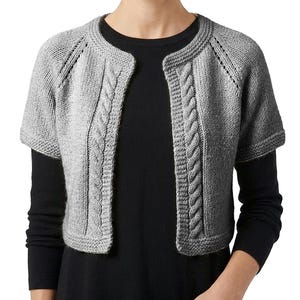 May include: A gray knitted bolero with short sleeves. The open front cardigan features a cable knit design along the edges and sleeves. The bolero is made of a soft, textured yarn and is worn over a black top.