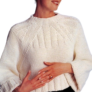 Könnte beinhalten: Ein weißer Strickpullover mit hohem Kragen und langen Ärmeln. Der Pullover hat ein Zopfmuster und eine lockere Passform.